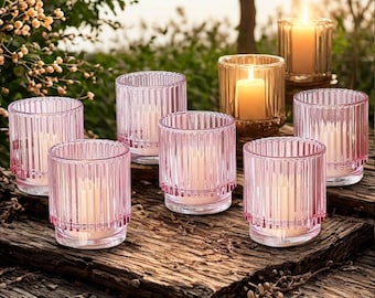 May include: A collection of glass candle holders in shades of pink and amber, arranged on a rustic wooden surface. The candle holders have a ribbed design and contain lit candles, creating a warm, inviting ambiance. The scene is set outdoors with natural greenery in the background.
