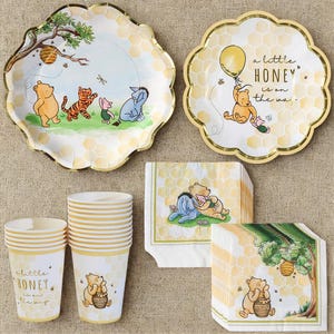 May include: A collection of Winnie the Pooh themed party supplies. Includes plates, cups, and napkins with illustrations of Pooh, Tigger, Piglet, and Eeyore. The plates and cups have a honeycomb pattern and gold trim. The text "a little HONEY is on the way" is printed on the cups and plates.