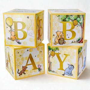 May include: Four yellow baby blocks spelling "BABY". Each block features a letter, illustrations of Winnie the Pooh characters, and a honeycomb pattern. The blocks are stacked, creating a playful and decorative arrangement.