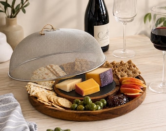 May include: A round wooden serving tray featuring a tiered design with a smaller circular slate insert. A silver-toned metal mesh food cover with a small rope handle sits over crackers. The tray holds assorted cheeses, green olives, and jam.