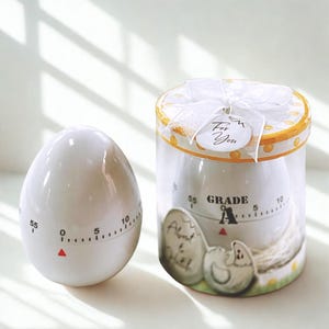 May include: White egg-shaped kitchen timer with a red indicator and a dial marked from 0 to 55. The timer is next to a clear cylindrical container holding a similar timer, decorated with egg-themed graphics and a "For You" tag.