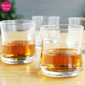 May include: Set of four clear, curved-bottom glasses, each holding amber liquid. The glasses are on a light-colored wooden surface. The image includes a pink circle with the text "Set of 4 10 oz".