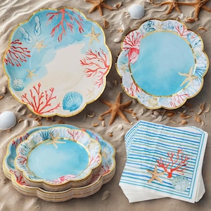 May include: A collection of beach-themed paper plates and napkins. The plates feature a scalloped edge with a gold trim, and are decorated with coral, starfish, and seashells. The napkins have blue and white stripes and matching coral and starfish designs.