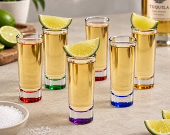 May include: A set of six clear glass shot glasses, each featuring a distinctively colored base in red, green, purple, blue, orange, and yellow. Each glass is filled with a golden liquid and topped with a fresh lime wedge, arranged on a surface.