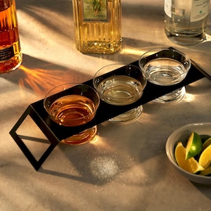 May include: A black metal serving tray holds three clear glass shot glasses, each filled with a different amber-colored liquid. A small bowl of lime wedges and a pile of salt are also present, suggesting a tequila tasting setup.