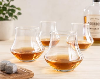 May include: Four clear glass tumblers with a wide base and tapered rim, each containing amber-colored liquid. A bottle of bourbon whiskey stands in the background, and two grey stone cubes rest on a small wooden board in the foreground.