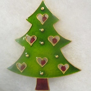 May include: A green Christmas tree-shaped brooch with gold trim and red heart-shaped cutouts. The brooch has a glossy finish and small gold accents. The base of the tree is a deep red color. A festive holiday accessory.