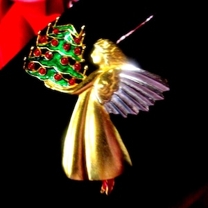 May include: A gold-toned angel brooch holding a green Christmas tree adorned with red ornaments. The angel has silver wings and a flowing gold dress. The brooch is set against a black background, highlighting the festive design.