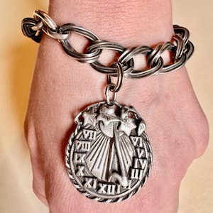 May include: Silver-toned bracelet with a large link chain design. A round pendant with Roman numerals and a sun dial design hangs from the bracelet. The bracelet is worn on a wrist.