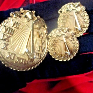 May include: A gold-colored jewelry set featuring a pendant and two matching earrings. The pieces are circular, with Roman numerals around the edge and a sun dial design in the center. The set has a textured, rope-like border and decorative leaves.