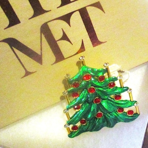 May include: A green Christmas tree brooch with red jewel ornaments and gold accents. The brooch is set against a beige background with the letters "MET" visible. A festive holiday accessory.