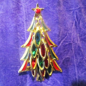 May include: A gold-toned Christmas tree brooch with red, green, and blue jewel-like accents. The tree is topped with a red star. The brooch is set against a purple background.