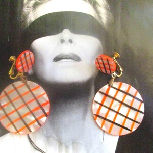 May include: Vintage clip-on earrings with a retro design. The earrings feature a round, translucent base with a grid pattern in orange and black. The smaller top piece is also orange with a black grid. The earrings have gold-colored clips.