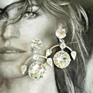 May include: A pair of silver-toned drop earrings featuring clear, faceted, round-cut crystals. Each earring consists of a large circular crystal suspended from a curved metal arch, accented with smaller round and pear-shaped crystals.