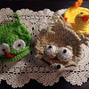 May include: Three crocheted coasters featuring character designs. One is a bright green creature with white eyes and a red tongue. Another is a tan creature with white eyes and a tuft of hair. The third is a yellow duck with orange wings and feet.
