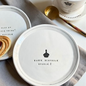 May include: White ceramic plate with a gray rim and the text "HAPPY BIRTHDAY STEVIE!" and a cake illustration. A cup of coffee and a gold spoon are in the background. A second plate with text is also visible.