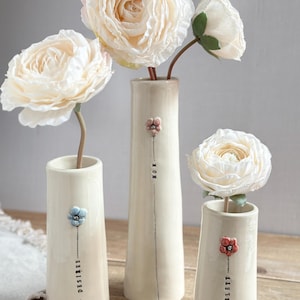 Peut inclure: Trois vases en céramique blanc cassé avec des fleurs et du texte. Les vases contiennent des fleurs blanches. Le texte sur les vases indique "DESIREE", "MOM" et "SCARLETT". Chaque vase a un accent floral de couleur différente.