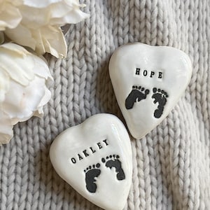 May include: Two white heart-shaped ceramic keepsakes with black text and footprints. One reads "HOPE" and the other "OAKLEY". The keepsakes are on a grey knitted surface, with white flowers in the upper left corner.