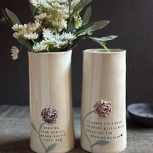 May include: Two cylindrical cream-colored ceramic vases featuring embossed pink flower appliques and black stamped text. The left vase reads "Ashlynn, grow gently, bloom boldly, love dad." The right vase reads "It takes a big heart to shape little minds, thank you for helping me grow."
