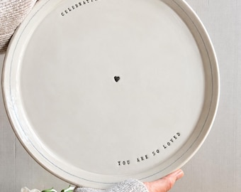 May include: A round white ceramic plate with a thin blue rim. The text "CELEBRATING YOU" is stamped at the top, "YOU ARE SO LOVED" at the bottom, and a small black heart is centered in the middle of the plate surface.