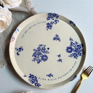 May include: Cream-colored ceramic plate with blue floral patterns and the text "YOU ARE SPECIAL TODAY!" and "KLINE FAMILY CELEBRATION PLATE". A gold fork is on the right side of the plate.