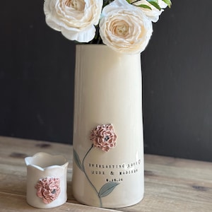 May include: A tall, cylindrical cream-colored ceramic vase and a small matching cup, both featuring a three-dimensional dusty pink flower appliqué. The vase displays the text "EVERLASTING LOVE JUDE & MADISON 8.28.26" with a painted green stem.