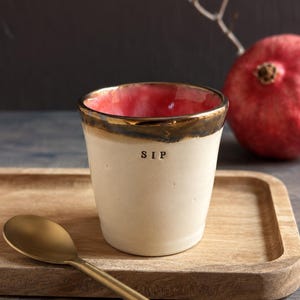 May include: A ceramic cup with a cream-colored exterior and a vibrant red interior. The rim is finished with a metallic gold glaze. The word SIP is stamped in black letters on the front. A matte gold spoon rests nearby on a wooden tray.