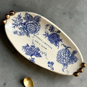 May include: An oval, cream-coloured ceramic tray with blue floral designs and gold accents. The tray features a blue rose and other floral patterns. The text "CELESTE + MICHAEL APRIL 13, 2026" is printed on the tray. A gold spoon is in the foreground.