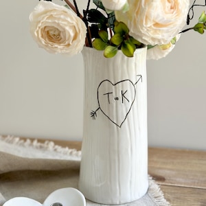 May include: A white ceramic vase with a textured bark-like surface, featuring a hand-drawn black heart pierced by an arrow with the letters T and K inside. Beside it sits a small white heart-shaped ceramic dish with the text BE MINE.