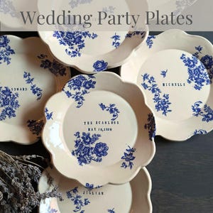 May include: A collection of white ceramic plates with a scalloped edge, adorned with blue floral patterns. Some plates feature names and dates in blue text, perfect for a wedding party. The plates are arranged on a dark surface.