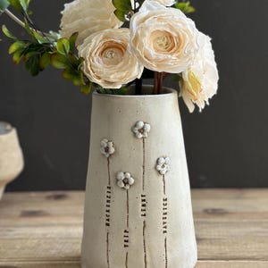 May include: A cylindrical off-white ceramic vase featuring four embossed flower stems. Each stem is labelled with the names Mackenzie, Trip, Lenny, and Maverick in black lettering. The vase is filled with a bouquet of cream-coloured artificial flowers.