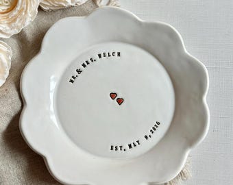 May include: A white ceramic flower-shaped dish featuring the black stamped text "MR. & MRS. WELCH" and "EST. MAY 9, 2026" along the inner rim, with two small red hearts centered in the middle of the plate, accompanied by a gold-toned metal fork.