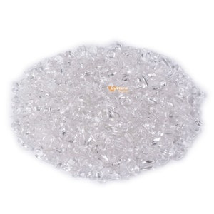 May include: A pile of clear quartz crystal chips. The crystals are small, irregular shapes, and appear translucent. The crystals are scattered across a white background, creating a sparkling effect. The crystals are likely used for jewelry making or other crafts.
