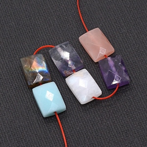 Pode incluir: Uma coleção de contas de pedras preciosas facetadas em forma retangular em várias cores, incluindo rosa, roxo, azul e branco, enfiadas em um cordão vermelho. As contas têm uma faceta em forma de diamante no centro. O fundo é cinza escuro.