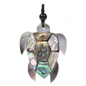 Puede incluir: Un colgante de tortuga marina hecho de concha de abulón iridiscente. La concha está cortada en un patrón de mosaico y engastada en resina negra. El colgante está unido a un cordón negro.