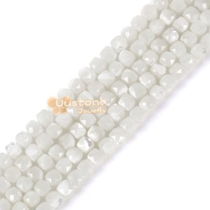 Puede incluir: Primer plano de un hilo de cuentas cuadradas blancas facetadas. Las cuentas tienen un aspecto ligeramente translúcido y están ensartadas. La imagen está bien iluminada, resaltando la textura y la forma de las cuentas. Para bisutería.