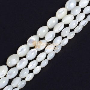 Könnte beinhalten: Eine Strähne aus weißen ovalen Muschelperlen. Die Perlen haben einen Perlmutt-Glanz und sind auf einem durchsichtigen Faden aufgefädelt.