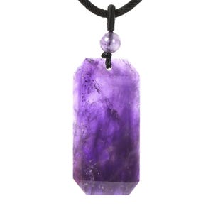 May include: A rectangular amethyst pendant with faceted edges suspended from a black cord. The stone features deep purple hues with natural internal inclusions and a small, round, polished amethyst bead positioned above the main pendant.
