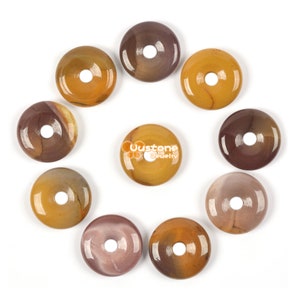 Puede incluir: Colección de piedras preciosas pulidas en forma de rosquilla, en tonos marrón, amarillo y beige, dispuestas en un patrón circular. Cada piedra tiene un agujero central. La piedra central muestra el texto "Uustone Jewelry".