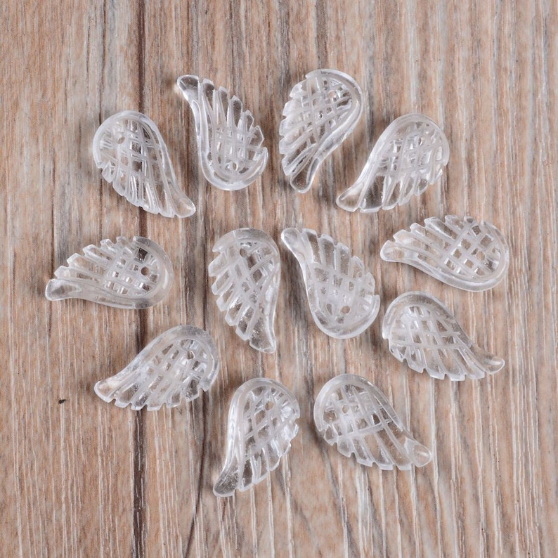 May include: A collection of twelve clear glass angel wing charms. The wings are arranged in a circle on a wooden surface.