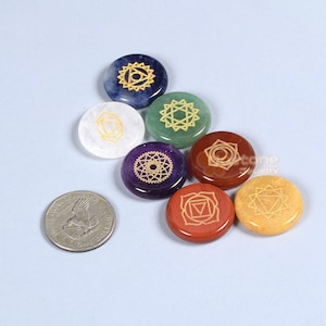 Może przedstawiać: Zbiór siedmiu kolorowych kamieni czakr ze złotymi symbolami, ułożonych na jasnoniebieskiej powierzchni. Kamienie są w różnych kolorach, w tym niebieskim, zielonym, białym, fioletowym, brązowym, czerwonym i żółtym. Obok kamieni znajduje się amerykańska moneta.