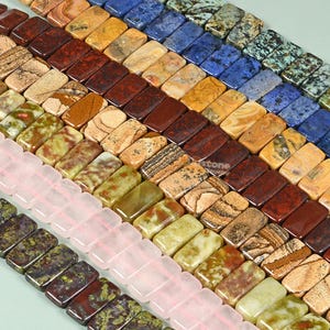 Könnte beinhalten: Sortiment an bunten, rechteckigen Edelsteinperlen. Die Perlen bestehen aus verschiedenen Steinsorten, darunter Jaspis, Achat und Quarz. Die Perlen sind in Reihen angeordnet, wobei die verschiedenen Farben und Muster ein optisch ansprechendes Display erzeugen.