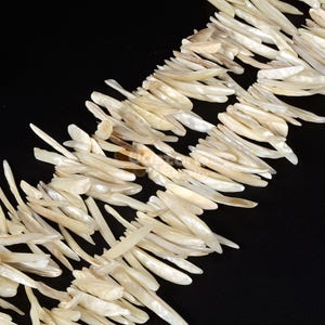 May include: A strand of white mother of pearl shell beads. The beads are long and narrow, resembling blades of grass. The beads are arranged in a random pattern, creating a natural and organic look.