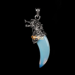 Könnte beinhalten: Ein silberner Wolfskopf-Anhänger mit einem blauen Opalzahn. Der Anhänger befindet sich auf einem schwarzen Hintergrund.