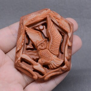 May include: A carved owl pendant made of red jasper. The owl is in flight and is surrounded by carved leaves. The pendant is a hexagonal shape.