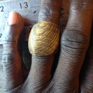 May include: A wide wooden ring featuring a prominent, swirling light and dark brown wood grain pattern. The ring is positioned next to a measuring tape showing a width of approximately 1.25 inches across the top surface of the band.