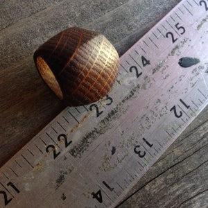 May include: A dark brown wooden ring with a visible grain pattern, positioned next to a metal ruler. The ring is aligned with the 23-inch mark on the ruler, showing a diameter of approximately 1 inch across the outer edge of the band.
