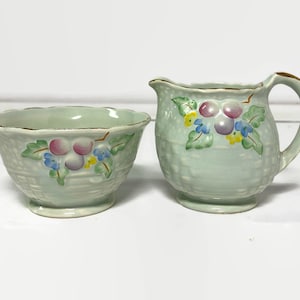May include: A vintage light green ceramic creamer and sugar bowl set. The set features a basket weave design with a floral pattern of pink, purple, blue, and yellow flowers. The rims are trimmed with gold.