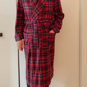 Op de afbeelding: Een lange, rood-zwarte geruite badjas met bijpassende riem en zakken. De badjas is gemaakt van een zacht, warm materiaal, perfect om in te loungen. De badjas wordt gedragen over een donker overhemd en een zwarte broek.