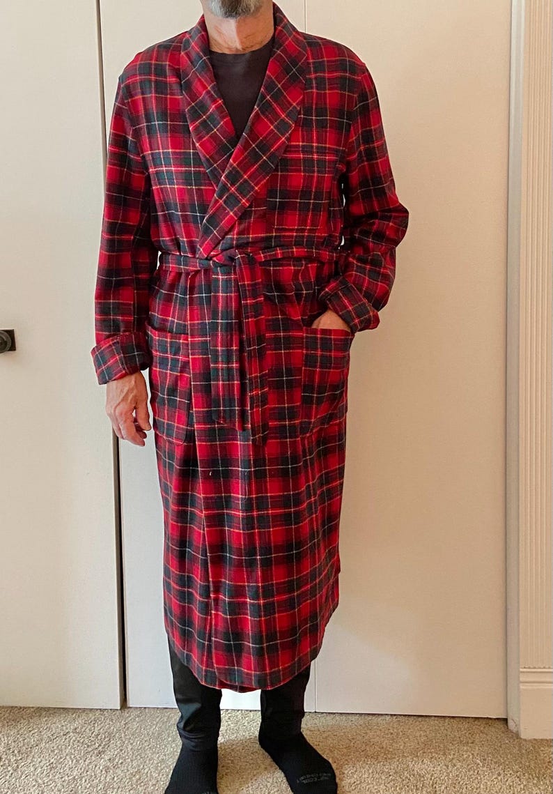 Op de afbeelding: Een lange, rood-zwarte geruite badjas met bijpassende riem en zakken. De badjas is gemaakt van een zacht, warm materiaal, perfect om in te loungen. De badjas wordt gedragen over een donker overhemd en een zwarte broek.